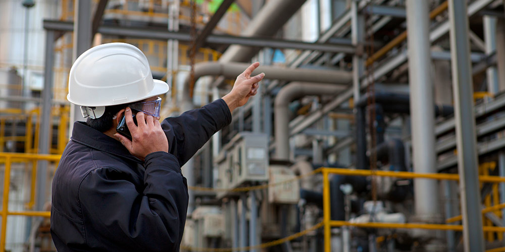 man with hard hat on cell phone pointing to metal piping in industrial facility