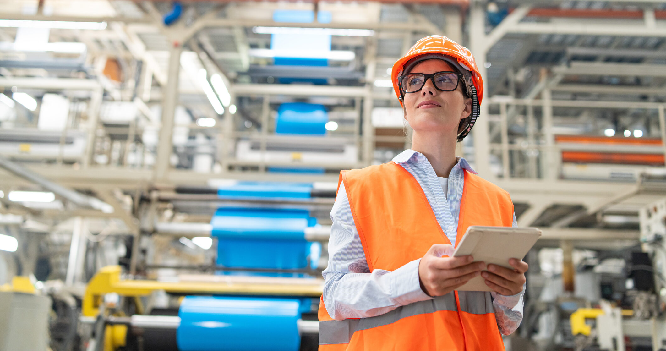 Engineer overseeing operations on tablet in bright industrial plant wearing safety gear. A female engineer wearing a bright orange vest and hard hat uses her tablet to oversee operations, embodying modern industrial oversight and safety protocols.